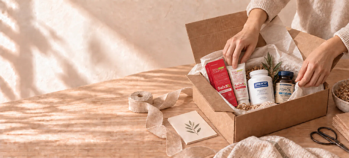 A person's hands are shown packing a curated box containing various wellness natural health products like a red and white tube, a jar of supplements, and a small bottle, set on a warm wooden table with soft natural shadows.